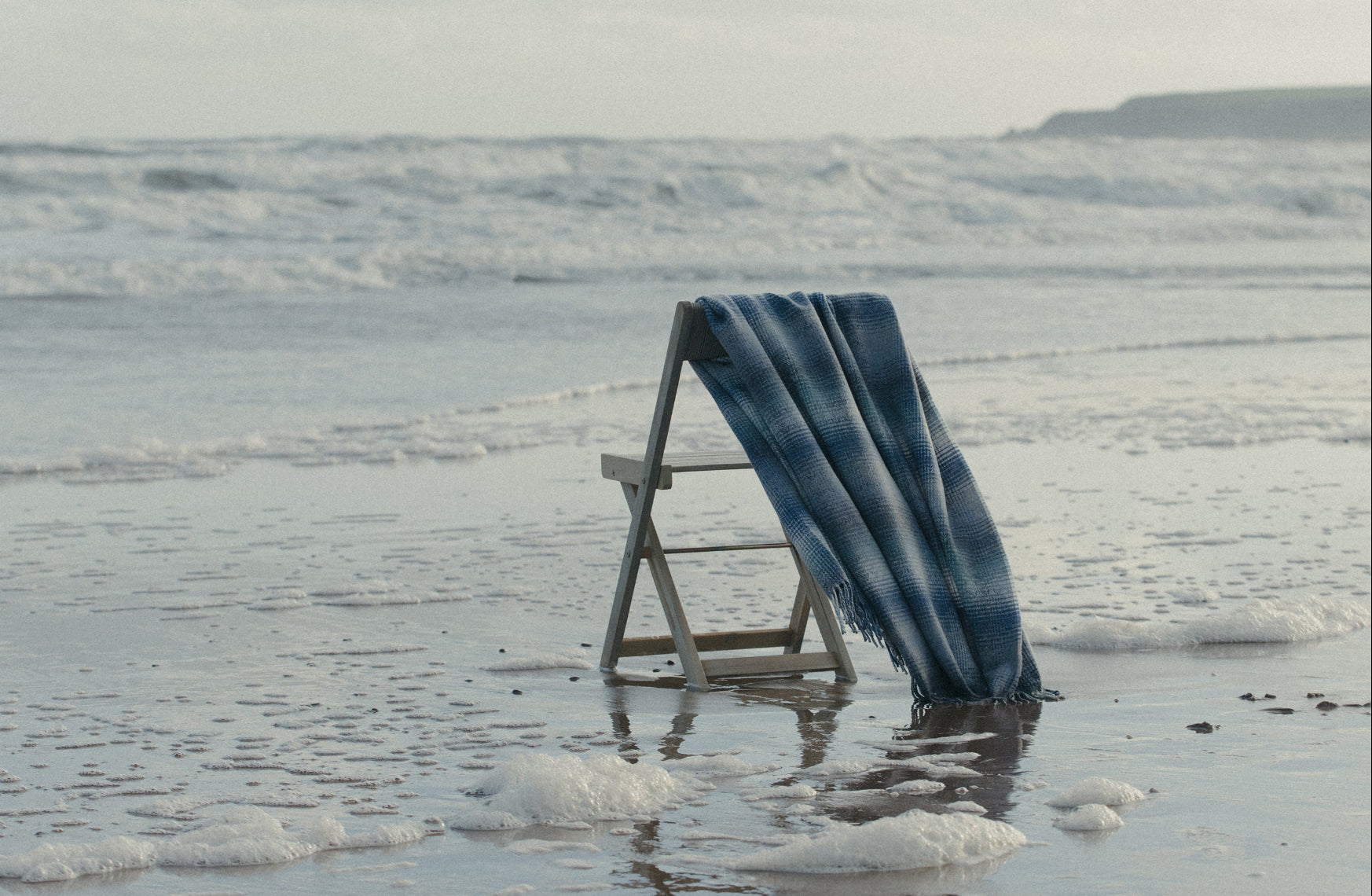 Blue Lunan Tidal Remony throw draped over a wooden chair on a beach with waves in the background