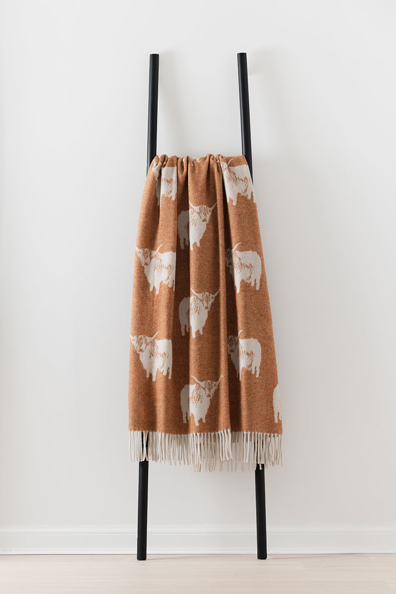 Remony nature highland cow rust throw - Brown blanket with white animal pattern draped over a black ladder against a white background