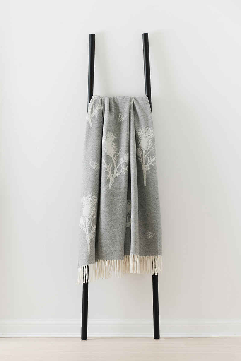Remony nature thistle and bee grey blanket with white pattern and fringes on a black stand against a white background