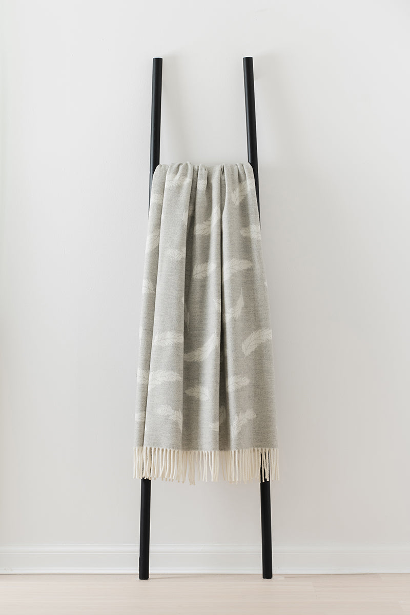 Remony Nature Feather silvery throw with white fringe draped over a black rack against a white background