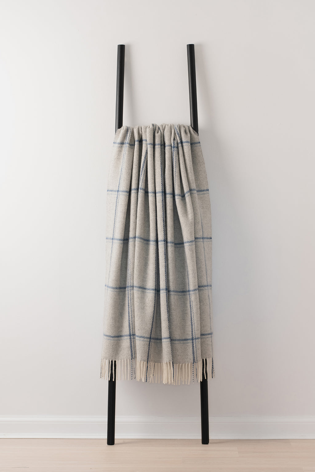 A grey-colored woven throw blanket with a blue windowpane plaid pattern and fringed edges, draped over a minimalist black metal ladder stand against a white wall.