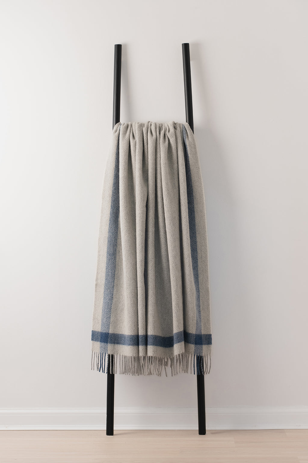A light grey woven throw blanket with blue stripes and fringe, draped over a minimalist black metal ladder stand against a white wall.