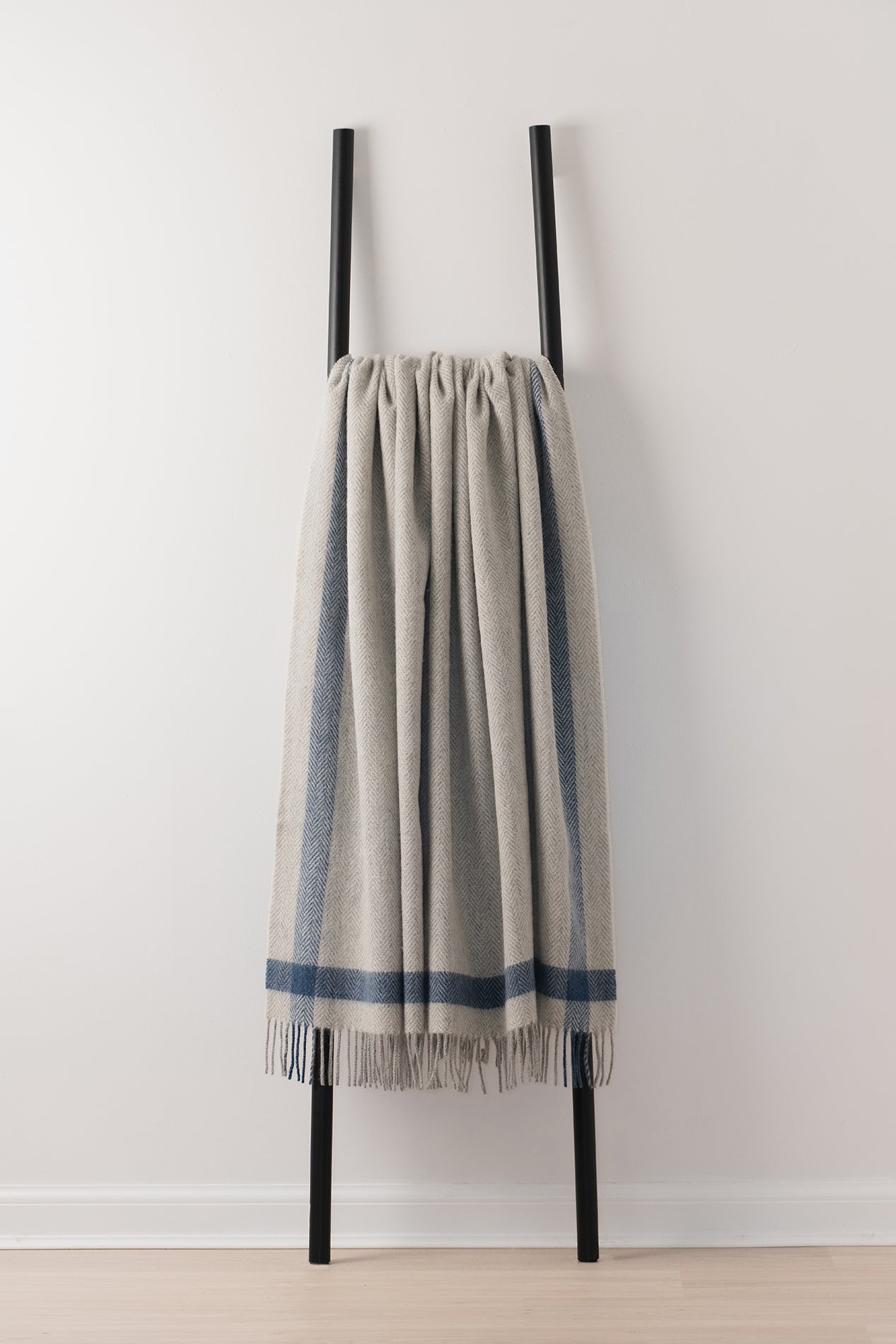 A light grey woven throw blanket with blue stripes and fringe, draped over a minimalist black metal ladder stand against a white wall.